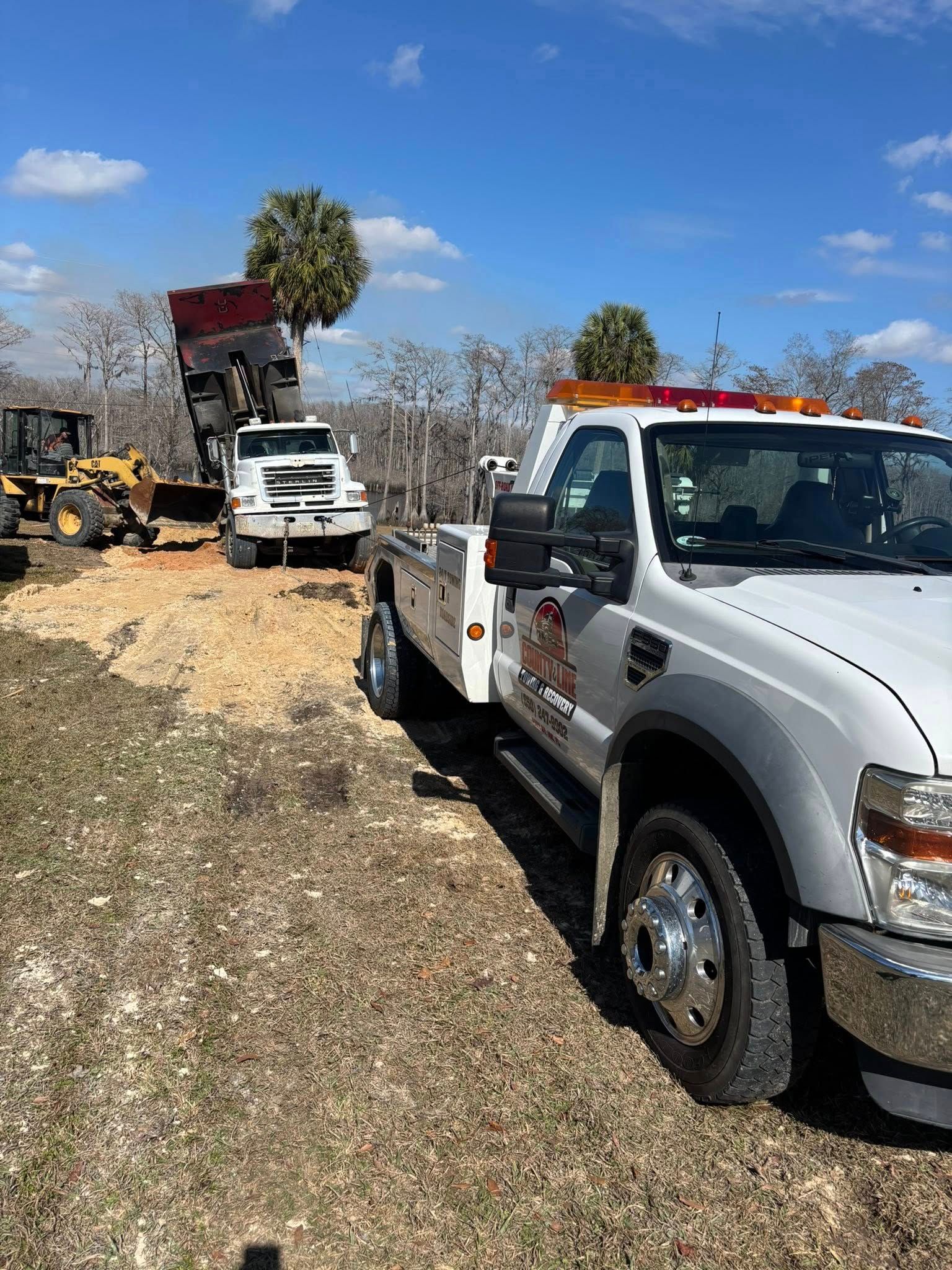 White tow truck parked; dump truck unloading dirt in a field under a blue sky.