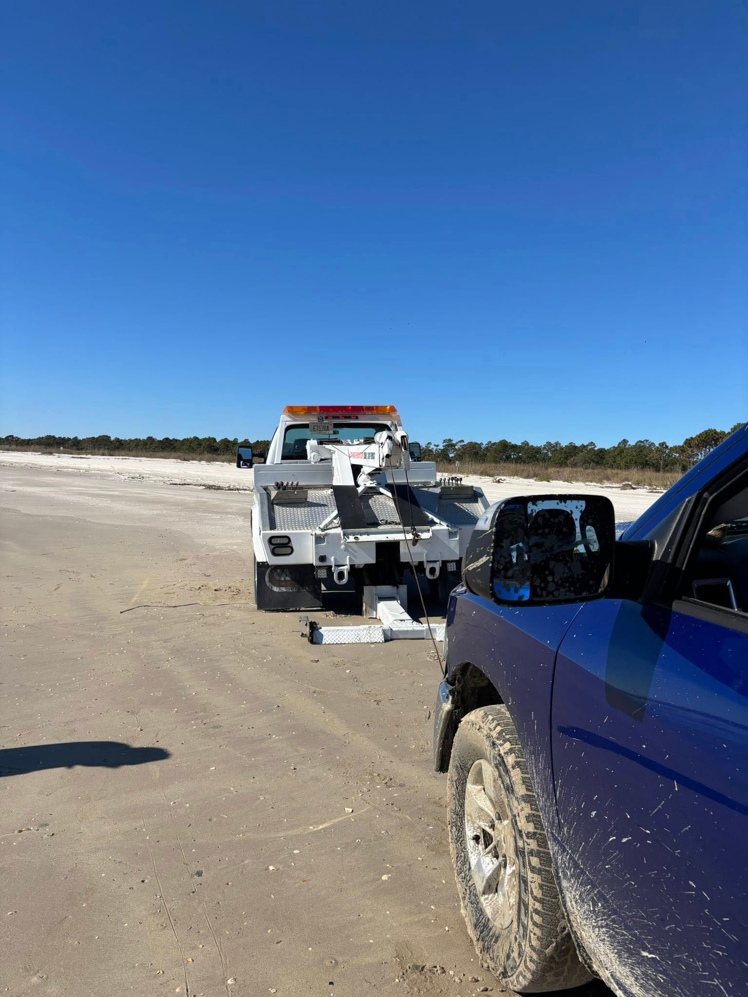 Tow truck towing a blue truck on a sandy beach under a clear, blue sky.