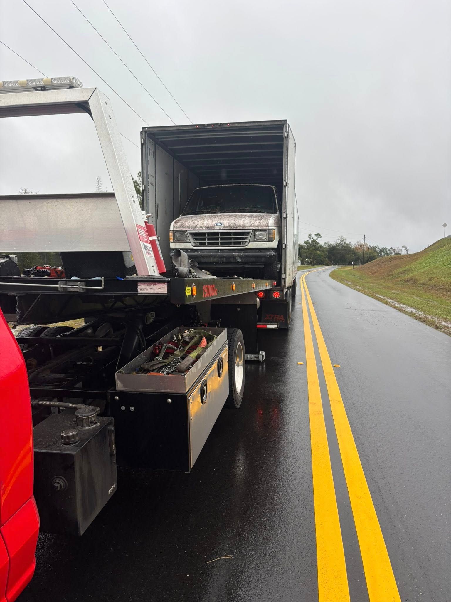 Truck loading a white pickup truck inside a trailer on a wet road.