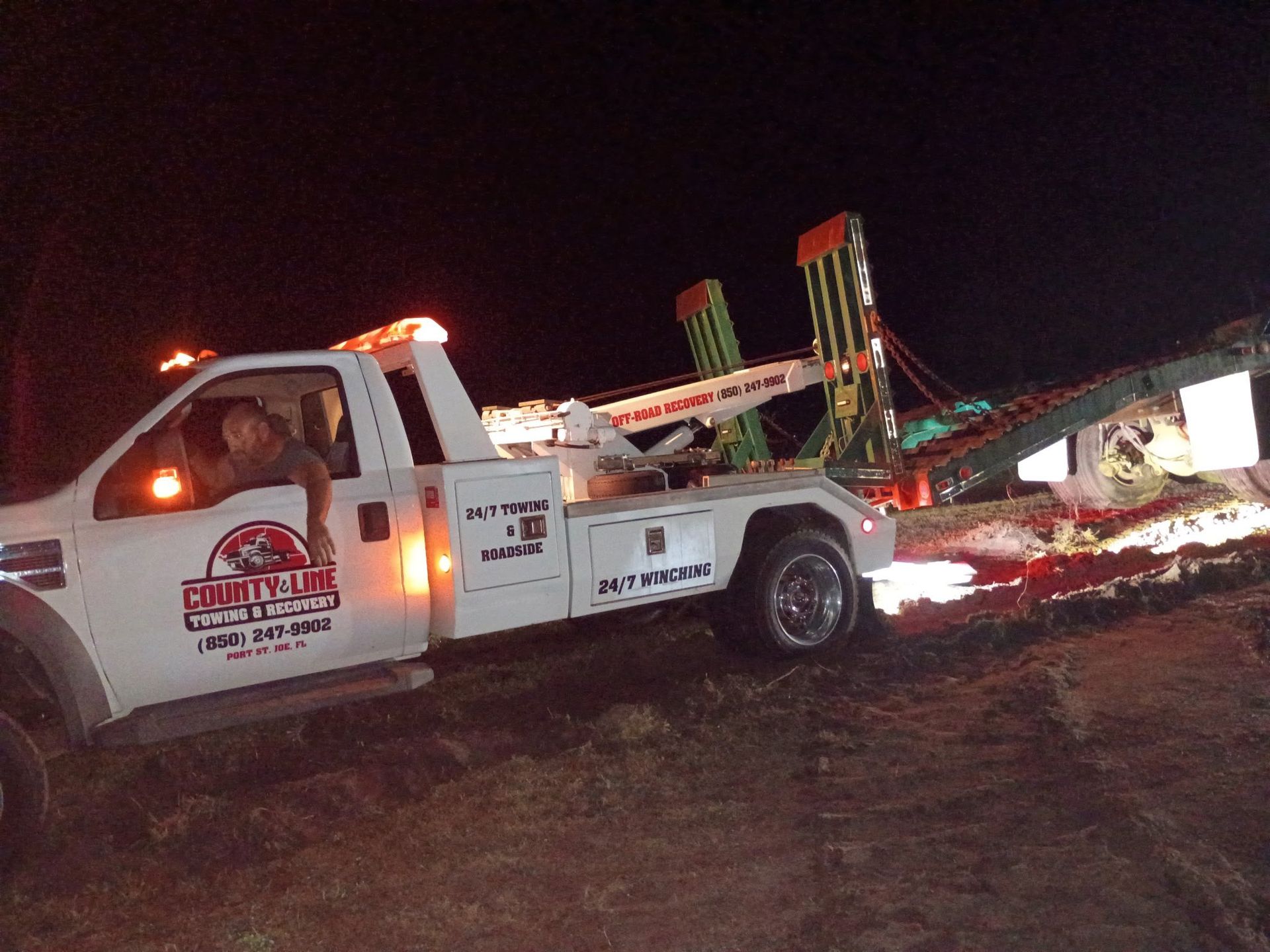 Tow truck on muddy ground at night, towing large green machinery.