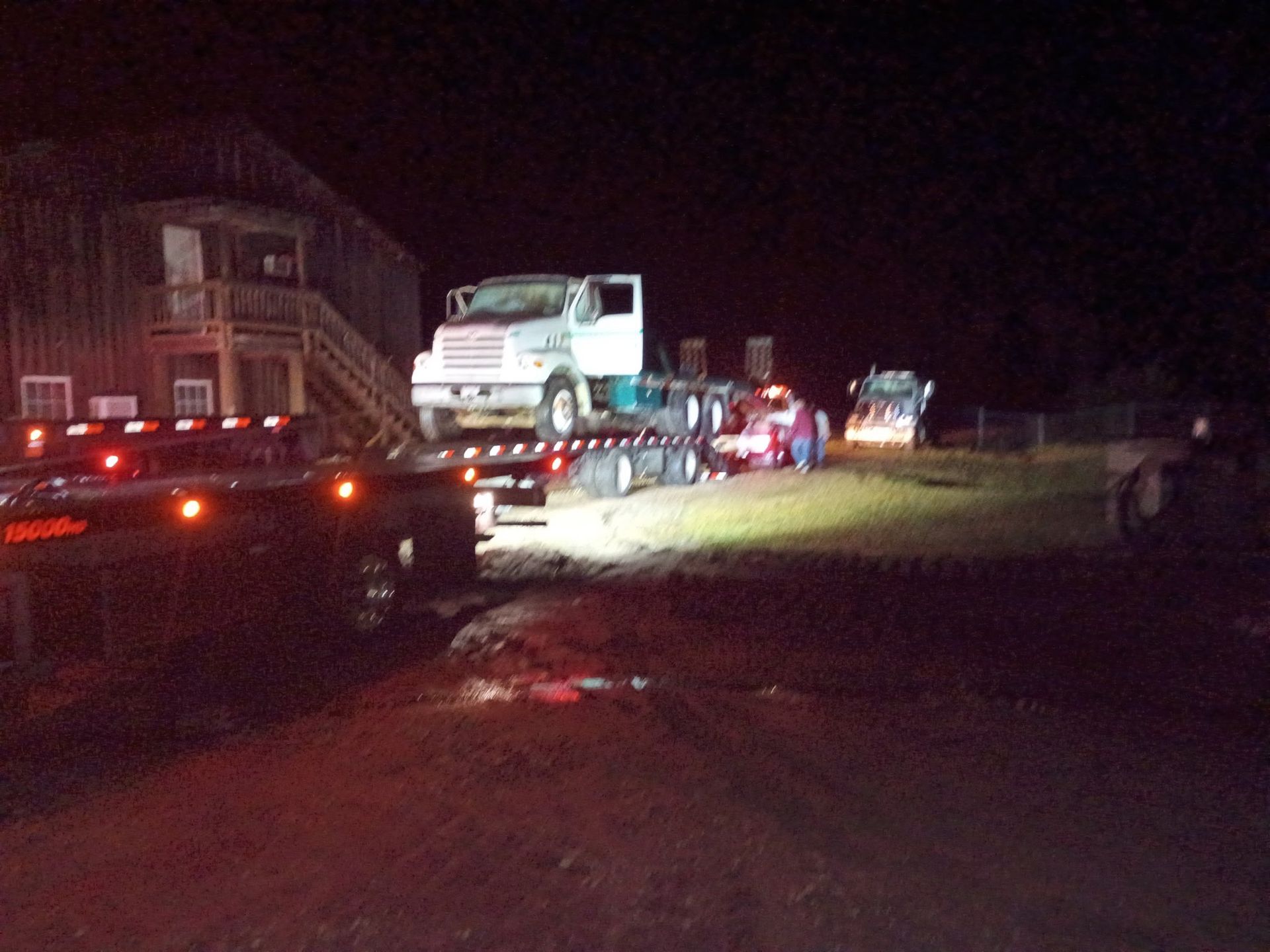 Trucks hauling vehicles at night. A truck transports a larger truck. Buildings in the background.