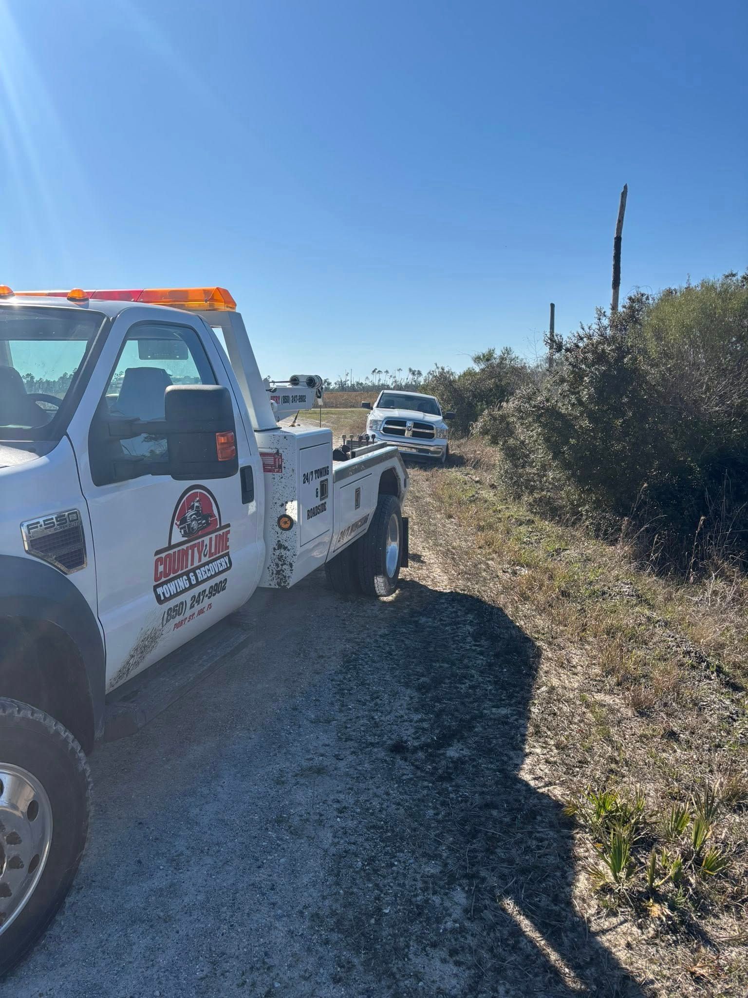 Tow truck towing a white vehicle on a dirt road next to vegetation under a blue sky.