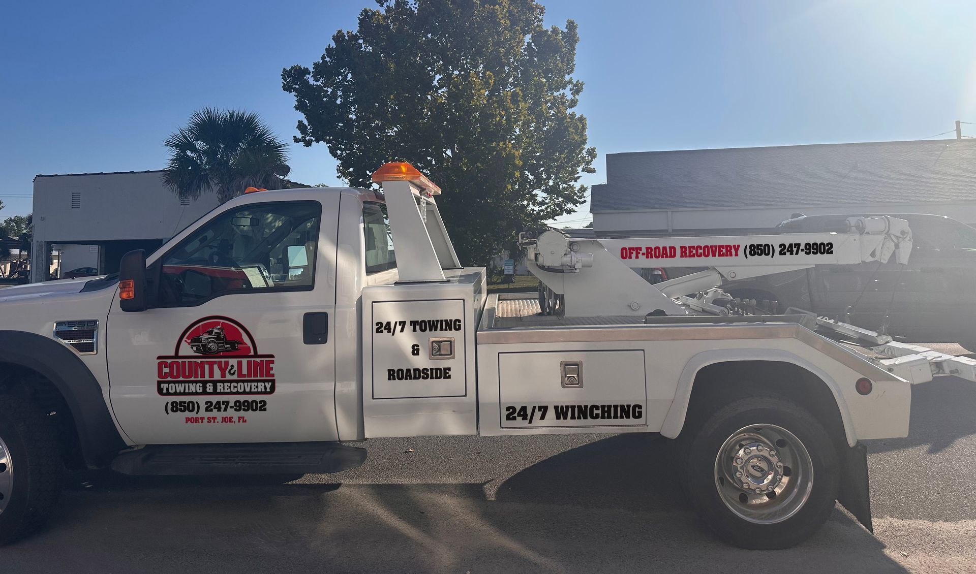 White tow truck with 24/7 service markings; parked outdoors under a sunny sky.