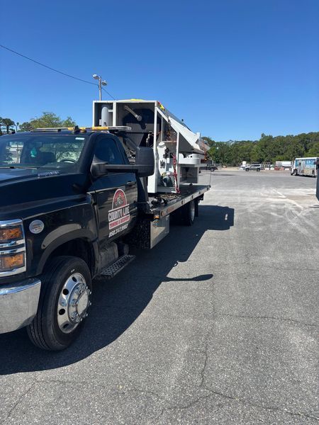 Black truck with white equipment on a flatbed trailer, parked on asphalt, sunny outdoor setting.