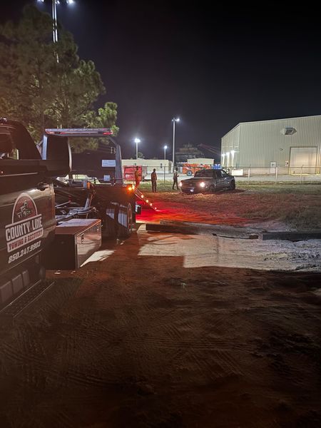 Tow truck at night, red lights flashing. A car is being loaded, near a building and people.