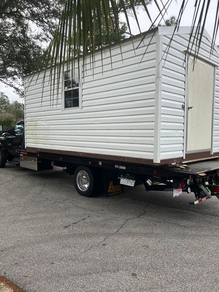 White shed loaded on a flatbed truck in an outdoor setting.