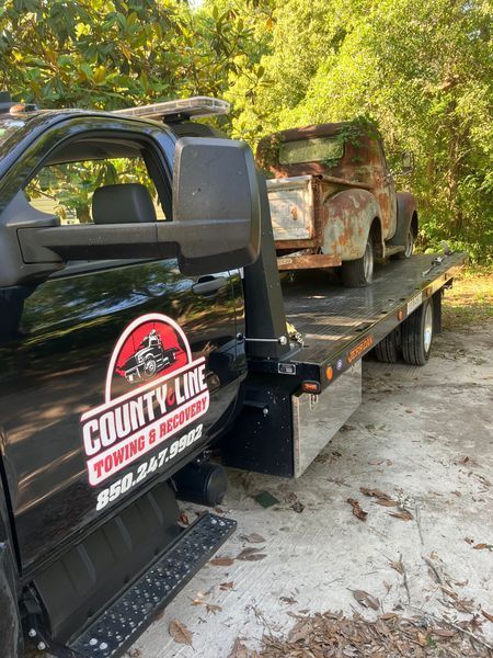 A rusted vintage pickup truck on a County Line Towing truck, outdoors.