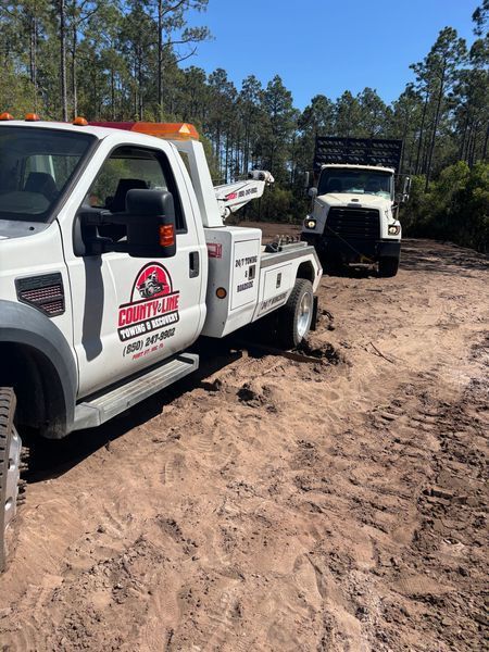 White tow truck pulling a large dump truck on a muddy road in a wooded area.