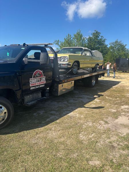 A classic yellow car loaded onto a black tow truck on a sunny day.