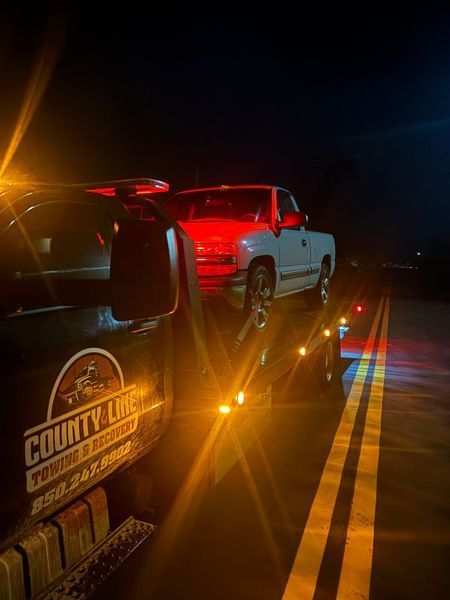 Tow truck hauling a white pickup truck down a dark road at night. The County Line Towing logo is visible.
