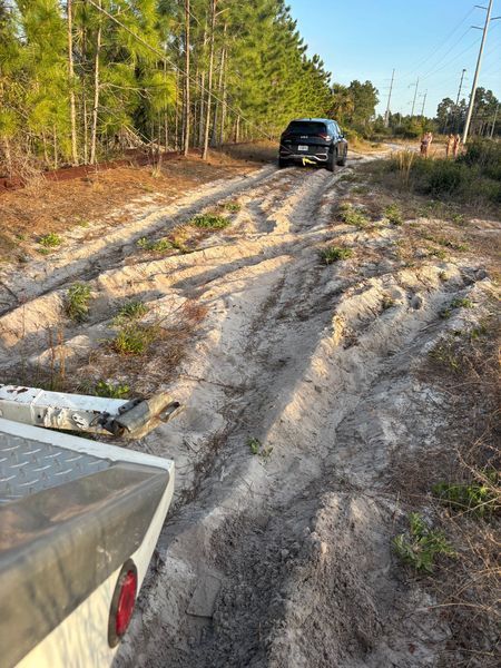 Black SUV on a sandy, rutted road. White trailer in the foreground. Trees in the background.
