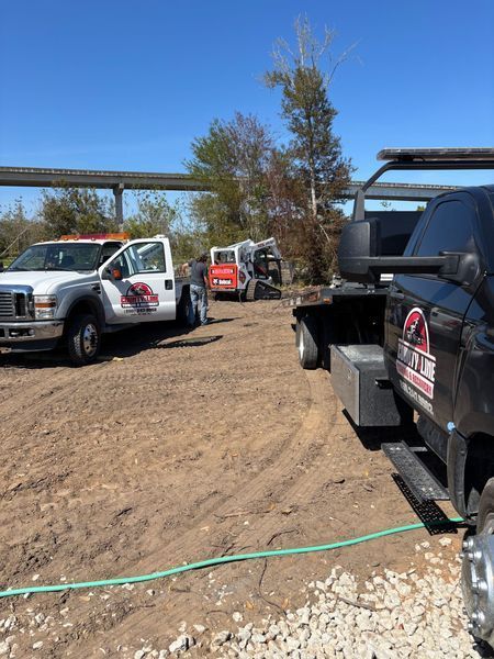 Construction vehicles parked on a dirt lot; two trucks, a Bobcat, and trees under a blue sky.