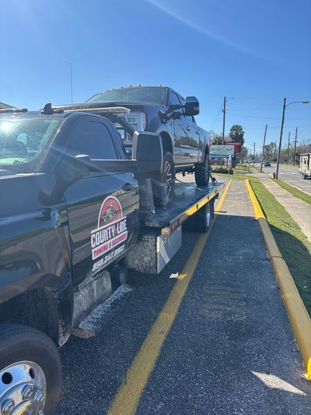 Black tow truck hauling a dark pickup truck on a sunny street.