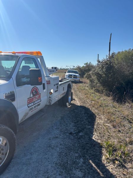 Tow truck towing a white car off a dirt road on a sunny day.