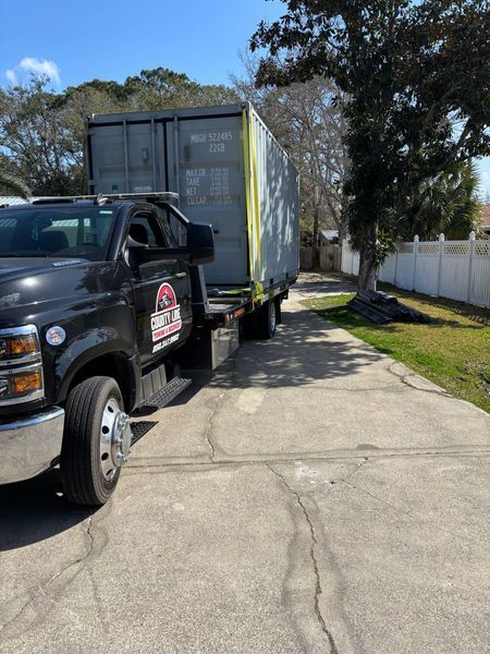 Black truck transporting a large gray shipping container down a paved driveway.