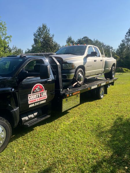 Silver Dodge truck being towed on a County Line Towing flatbed tow truck on grass under a clear sky.