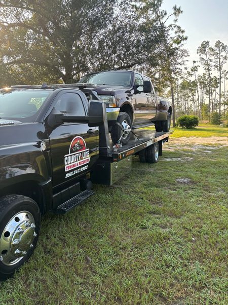 Tow truck hauling a dark-colored pickup truck on a grassy field, under a sunny sky.