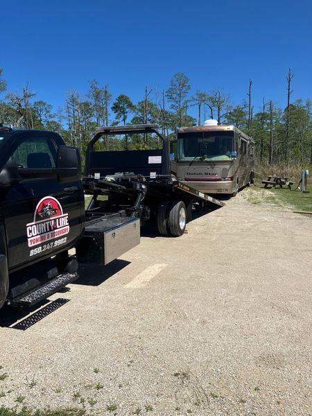 Tow truck towing a beige RV in a wooded area on a sunny day.