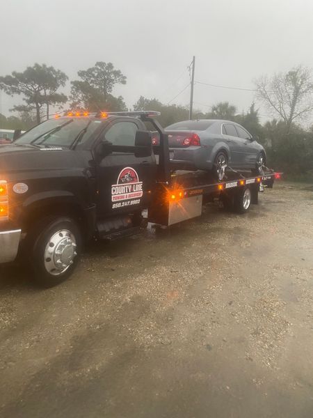 Black tow truck carrying a gray car on a wet, overcast day.