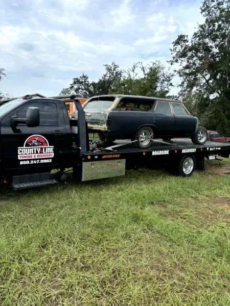 A black tow truck carries a dark blue station wagon on a flatbed. Outdoors, grassy field.
