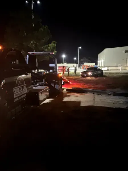 Tow truck towing a car at night; bright lights illuminate scene.
