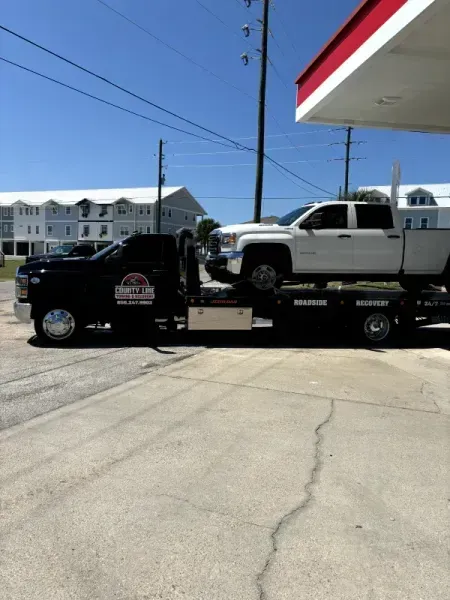 Black tow truck carrying a white pickup truck at a gas station under a sunny sky.