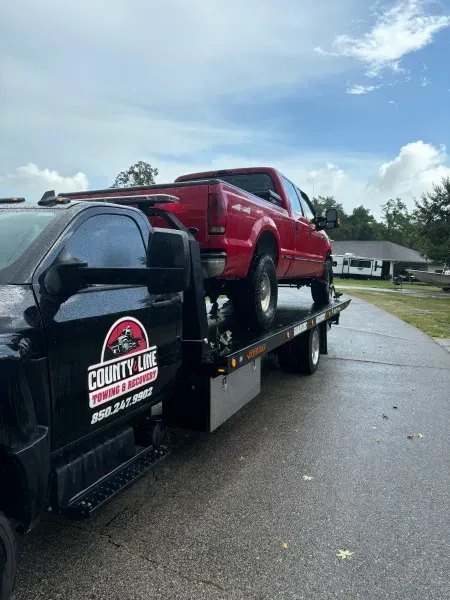Red pickup truck on a County Line Towing flatbed tow truck outdoors under a cloudy sky.