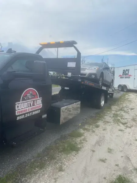 Tow truck with a car on its flatbed, parked on a gravel shoulder. The truck is black with the company name, County Line.