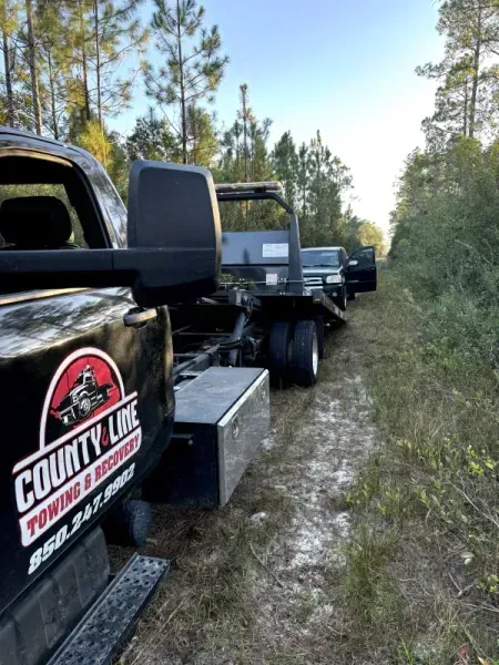 Tow truck, black and red logo, towing a vehicle on a dirt path, surrounded by trees.
