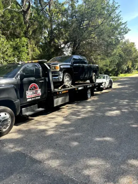 Tow truck carrying a black pickup truck and a white car on a road, trees in the background.