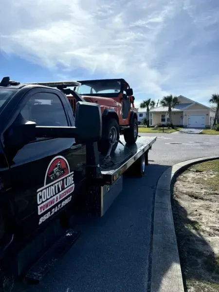 Tow truck hauling an orange Jeep on a sunny residential street. The truck is labeled 