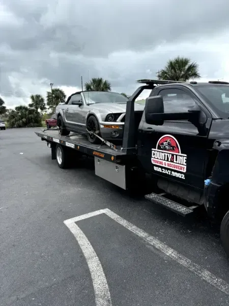 Silver convertible Mustang being towed by a County Line tow truck on a gray day.