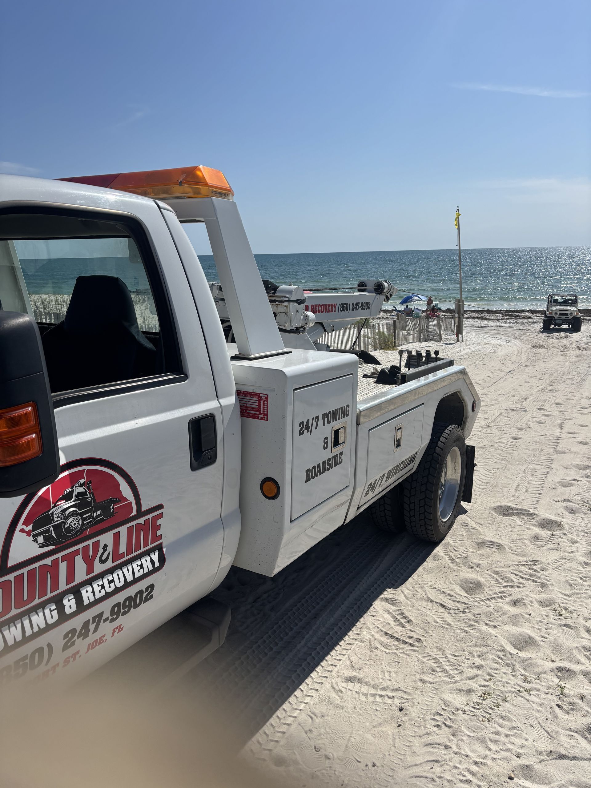 Tow truck on a sandy beach. Bright sunny day, blue water in background. 