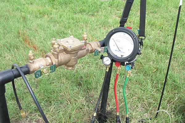 A brass water meter with gauges attached, set in green grass. Black pipes and supporting stand.
