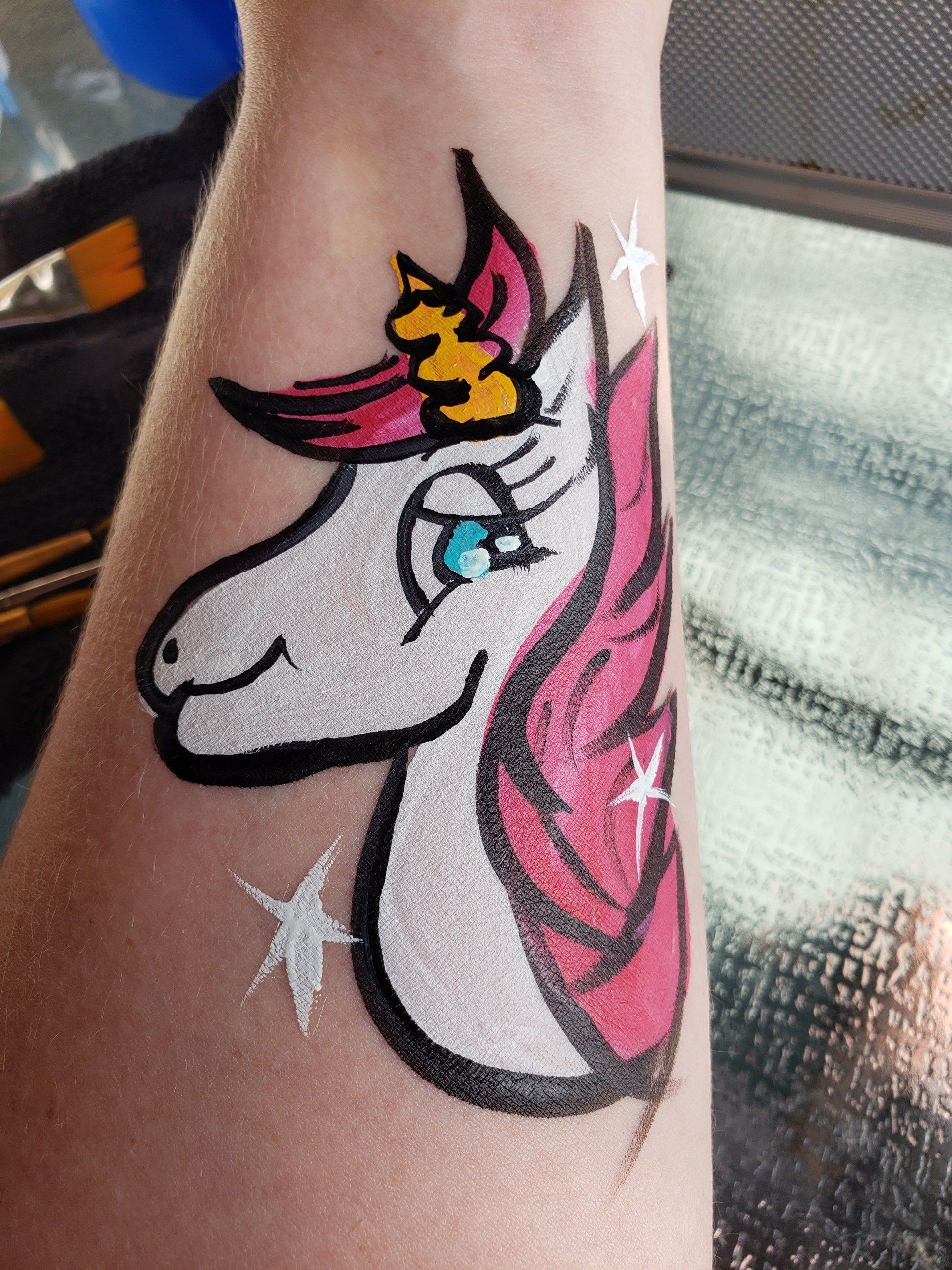 Unicorn with stars painted on inner forearm. Colors are white, pink, orange and teal for the eyes. 