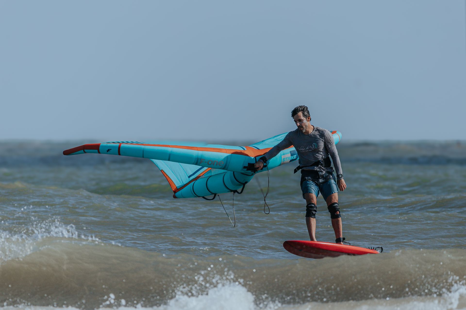Homem em uma prancha de foil, praticando wing surf sobre as ondas. Asa azul, prancha vermelha, cenário oceânico.