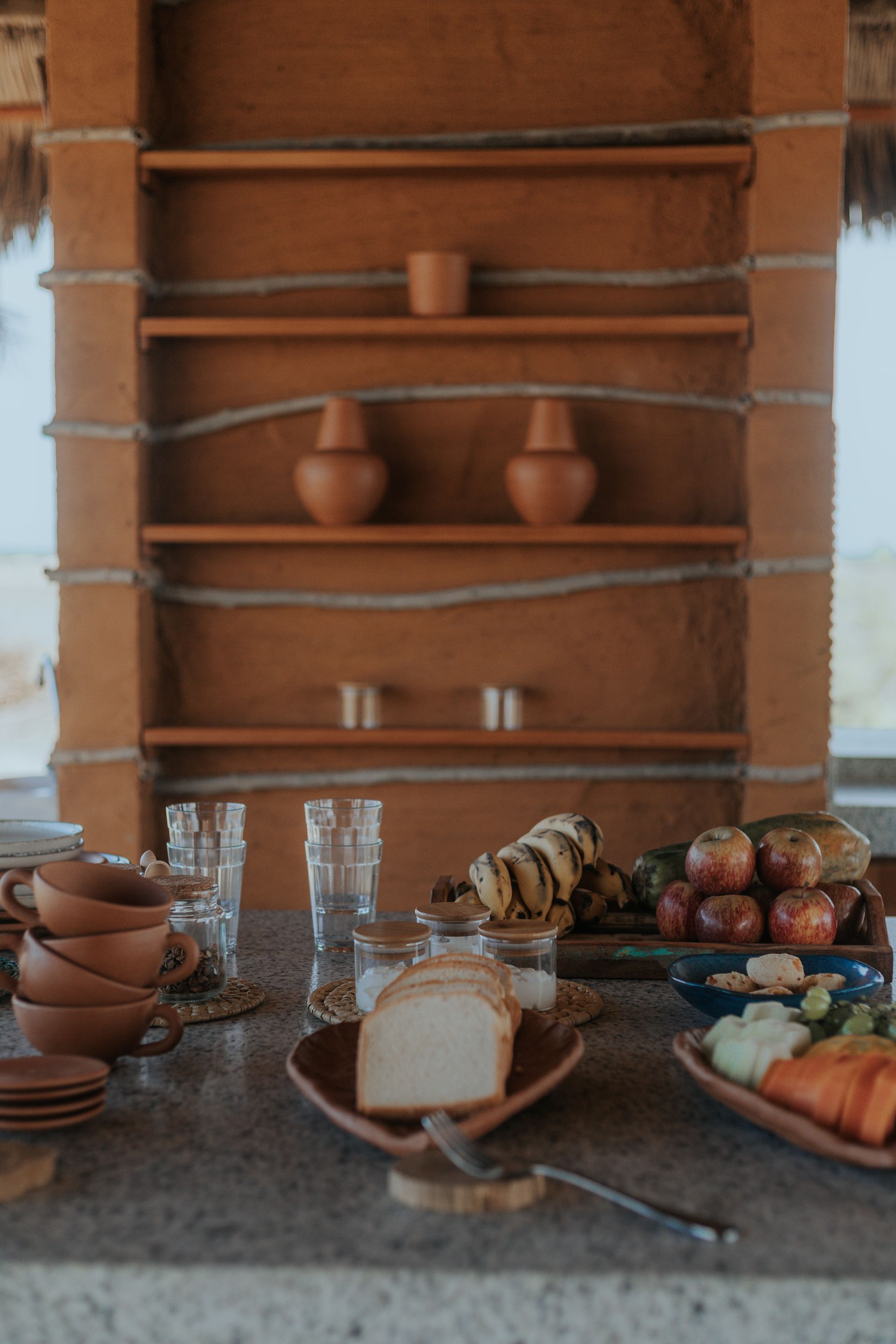 Bancada com alimentos, tigelas de cerâmica, copos e uma prateleira em tons terrosos com vasos.