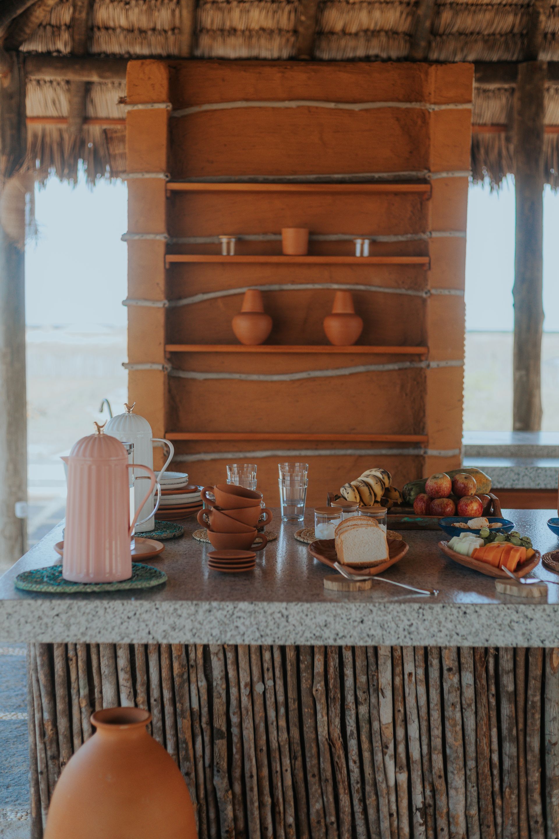 Cozinha rústica ao ar livre com prateleiras de barro, diversos utensílios de cerâmica, copos e alimentos.