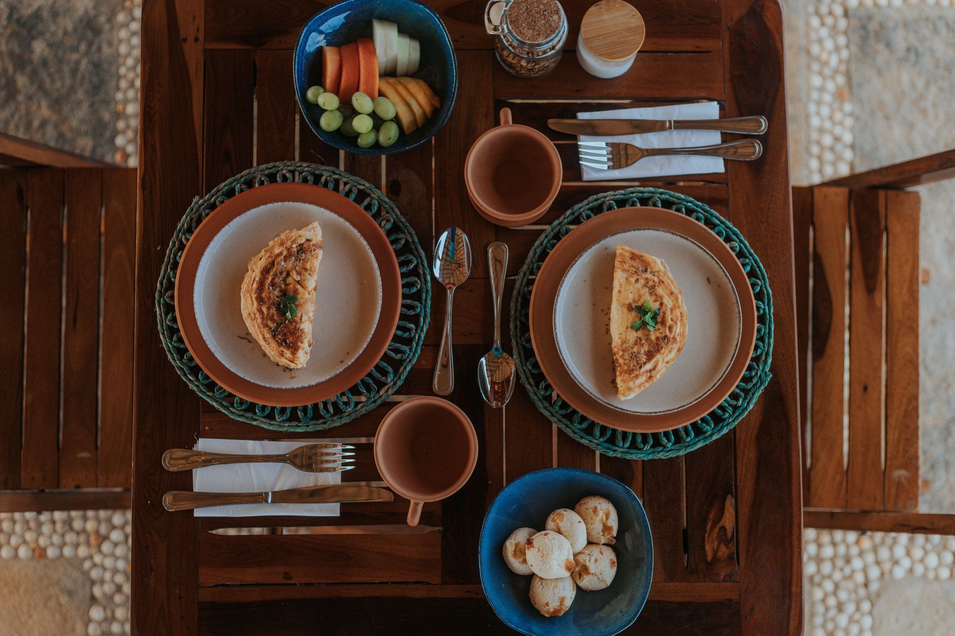 Mesa de café da manhã posta com pratos de comida, frutas e xícaras de café.
