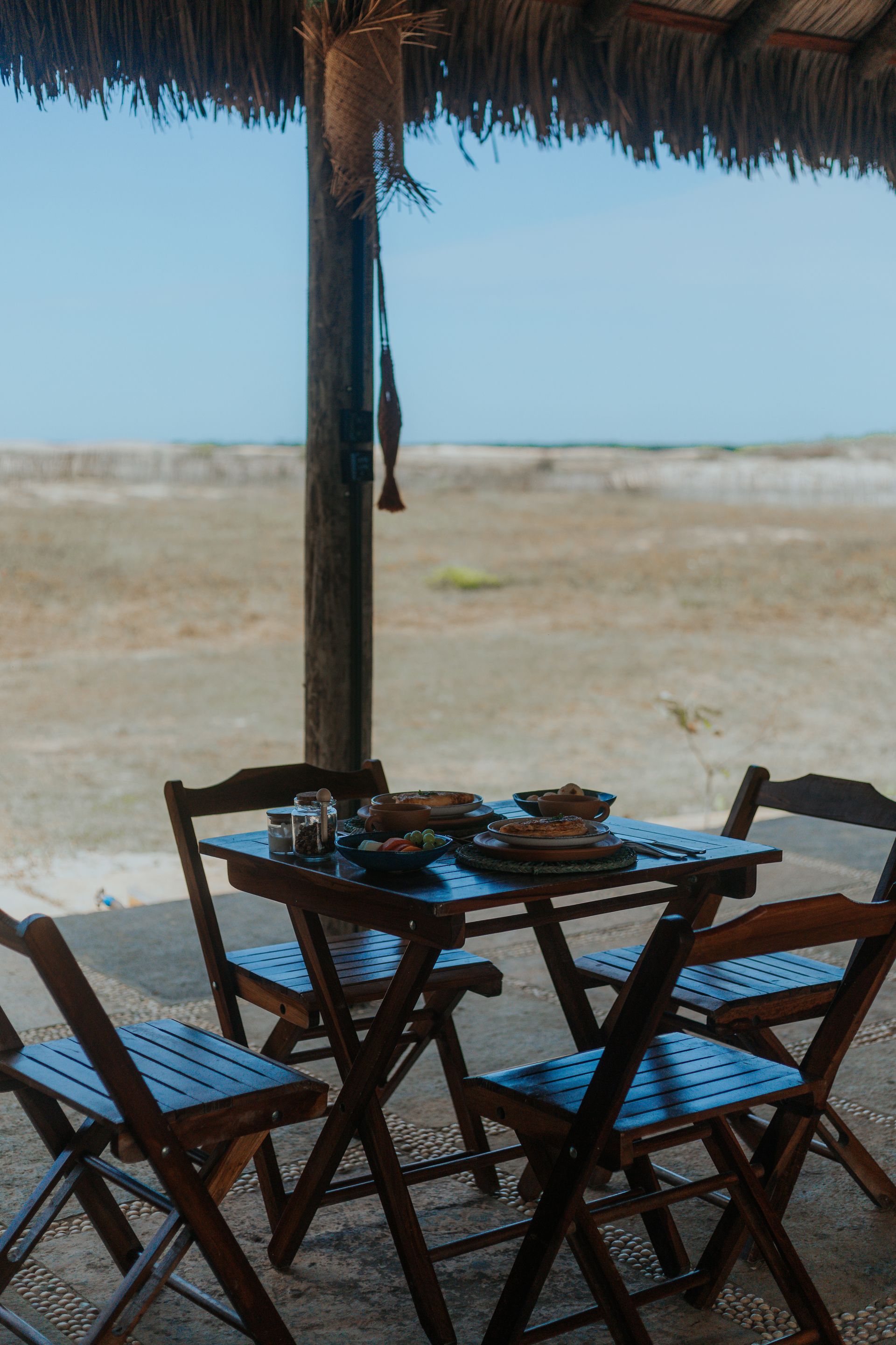 Uma mesa posta com comida e quatro cadeiras de madeira sob um telhado de palha, com vista para a praia.