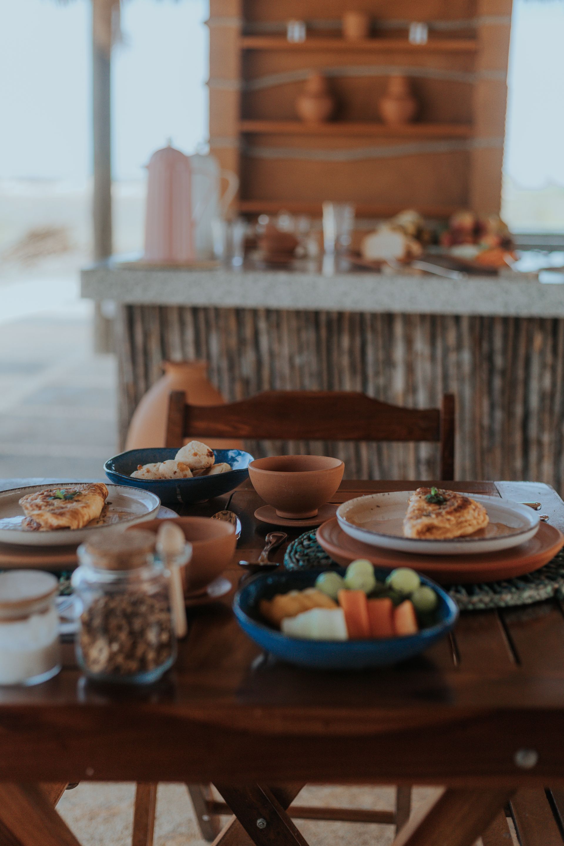Mesa de café da manhã posta com pratos de comida, frutas e cereais ao ar livre.