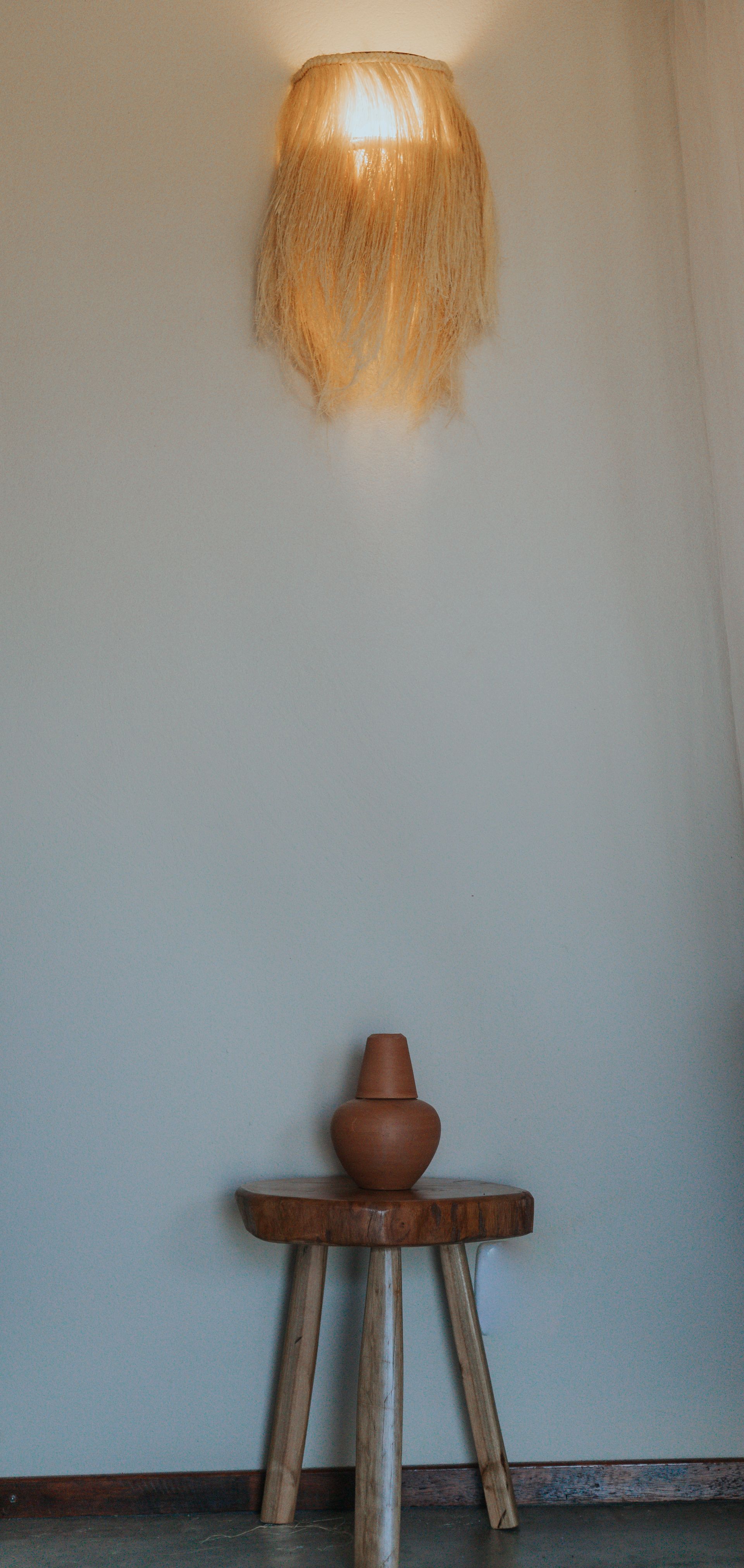 Um pequeno banco de madeira com um vaso de terracota sob uma luminária decorativa na parede.