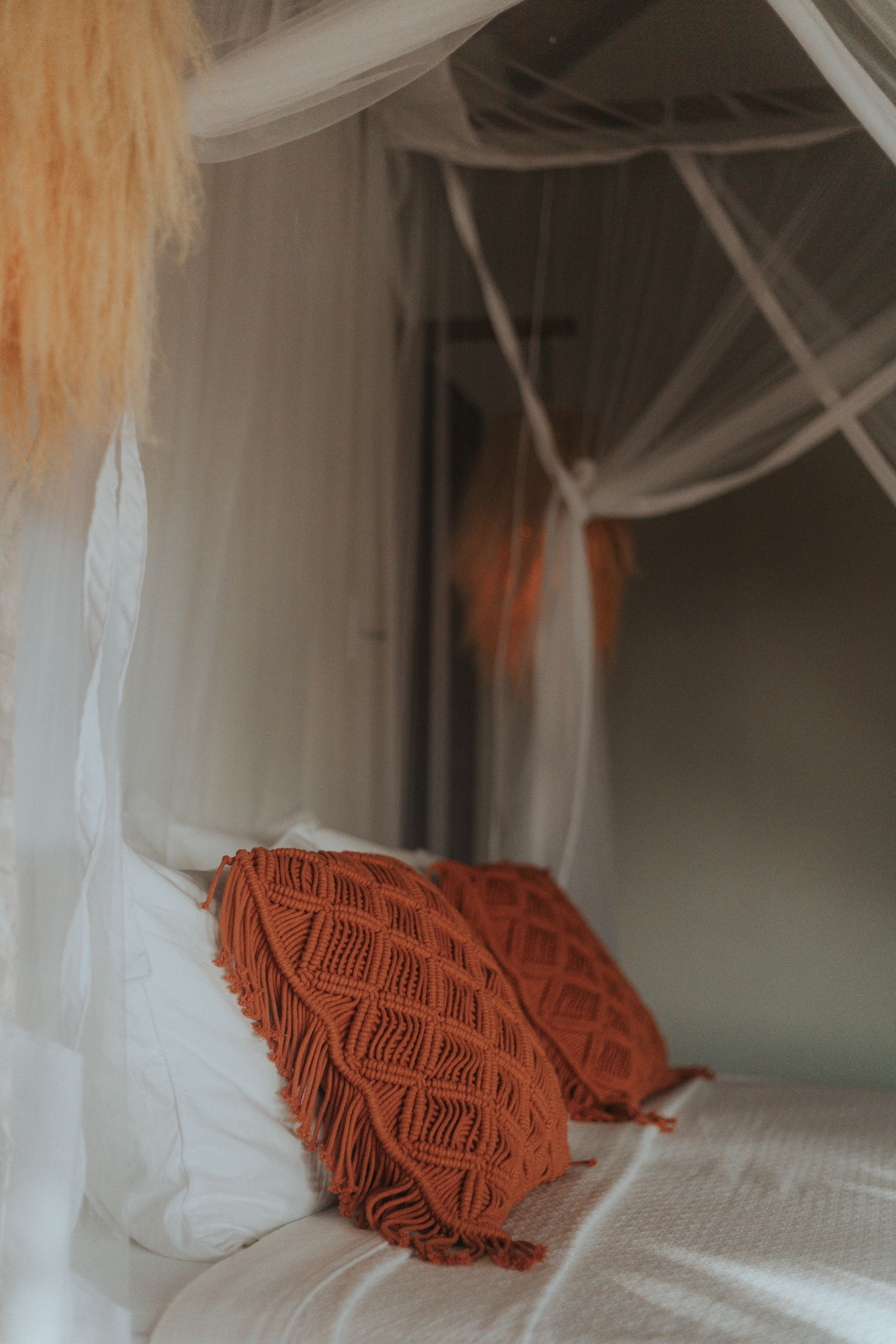 Cama com lençóis brancos, duas almofadas decorativas laranja e um dossel transparente.