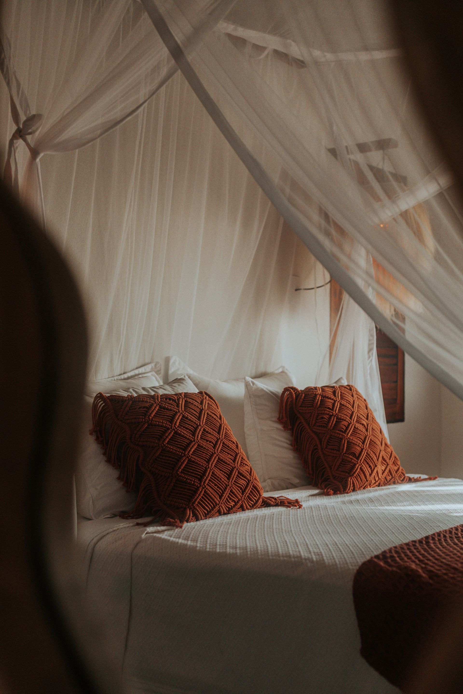 Cama com lençóis brancos, duas almofadas decorativas cor de ferrugem e um dossel de mosquiteiro.