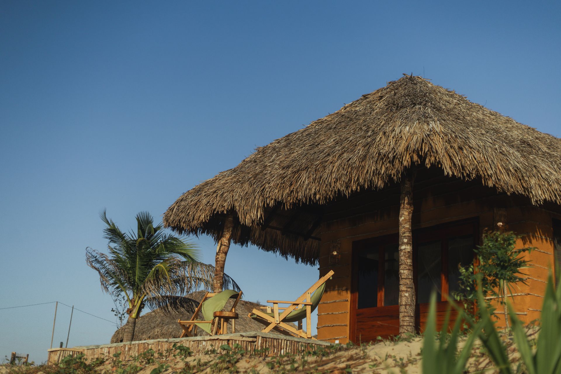 Cabana com teto de palha, postes de madeira, palmeira e céu azul.