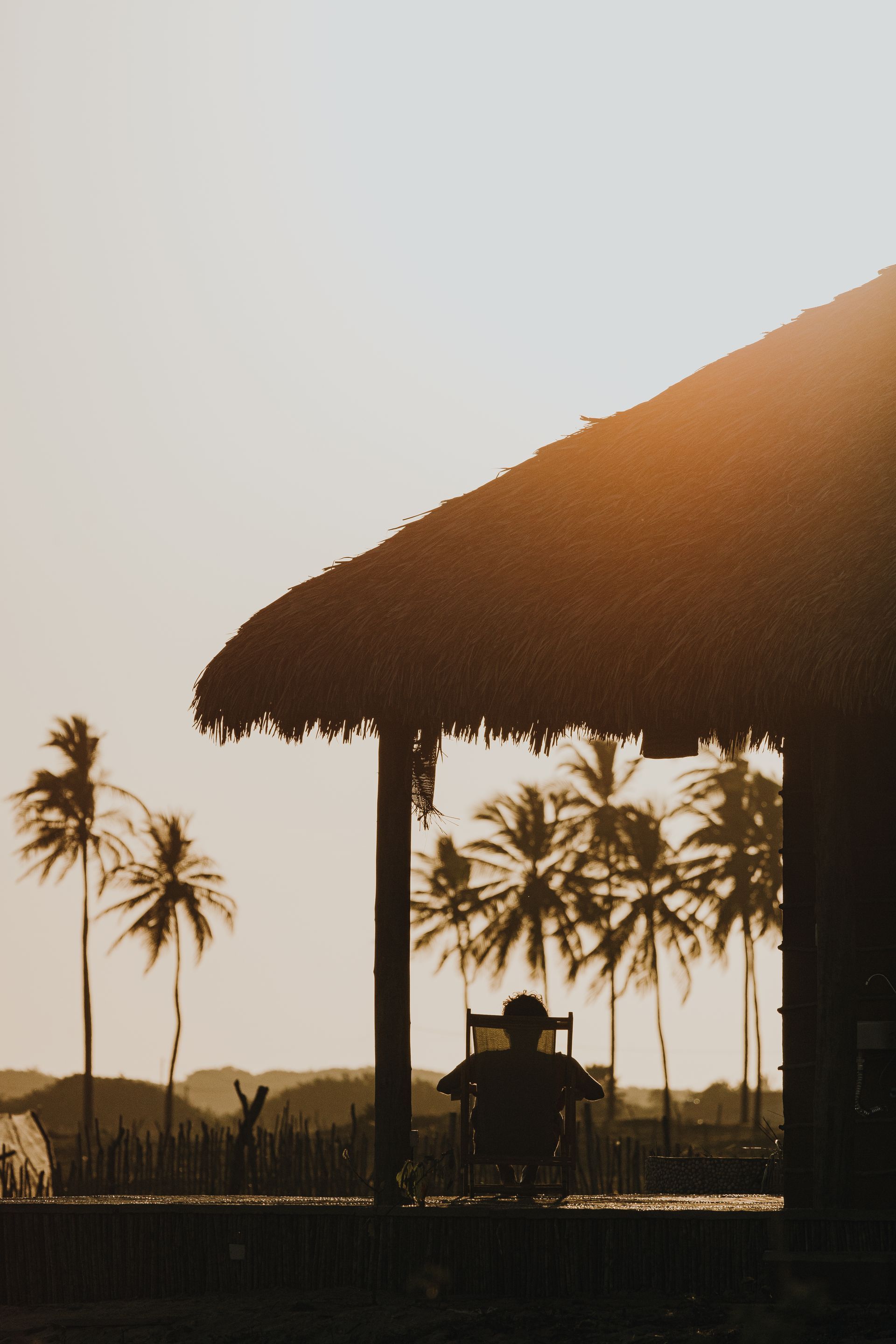 Silhueta de um abrigo com telhado de palha ao pôr do sol, com palmeiras ao fundo.