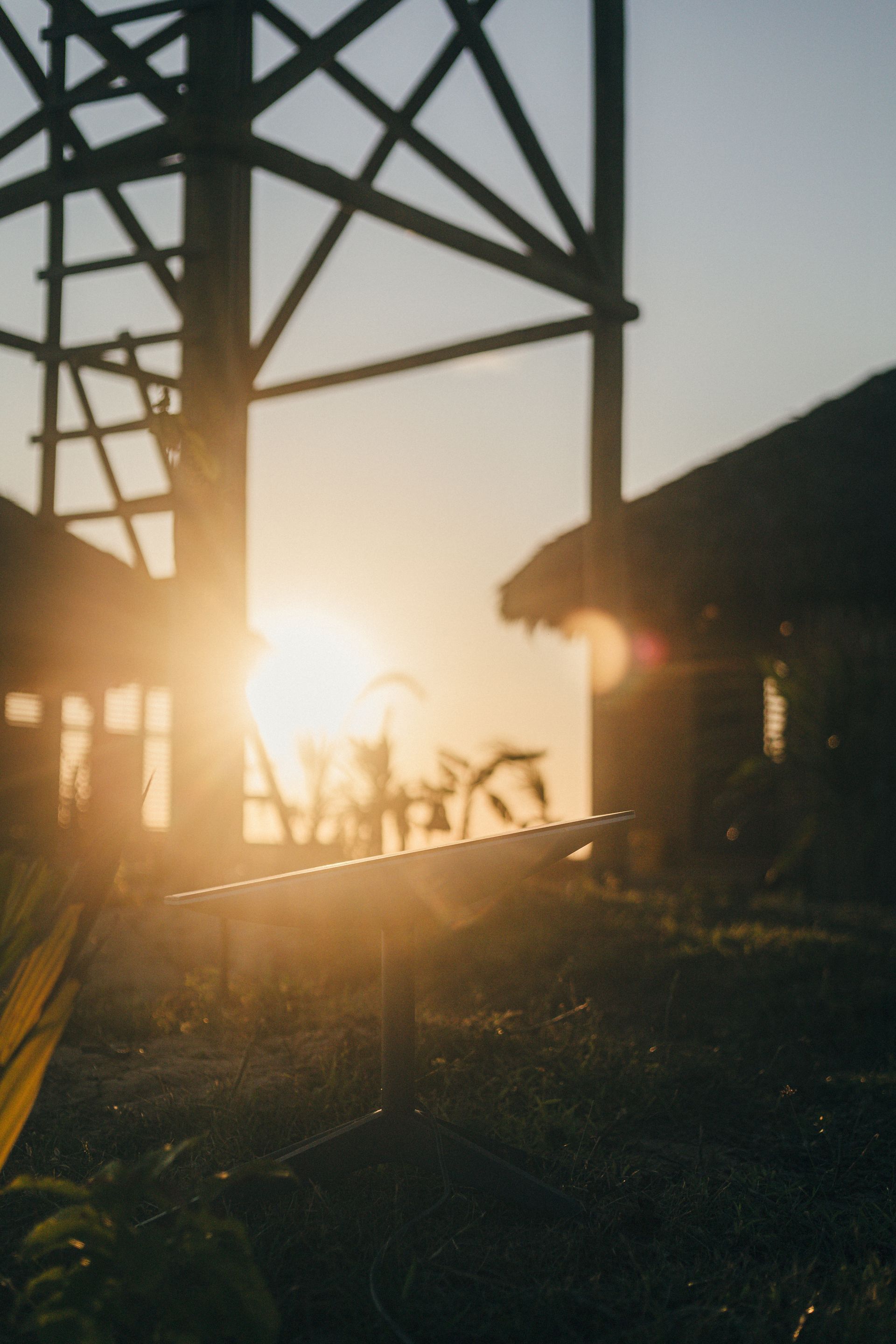 Uma antena Starlink na grama, em silhueta contra o pôr do sol, com estruturas de madeira ao fundo.