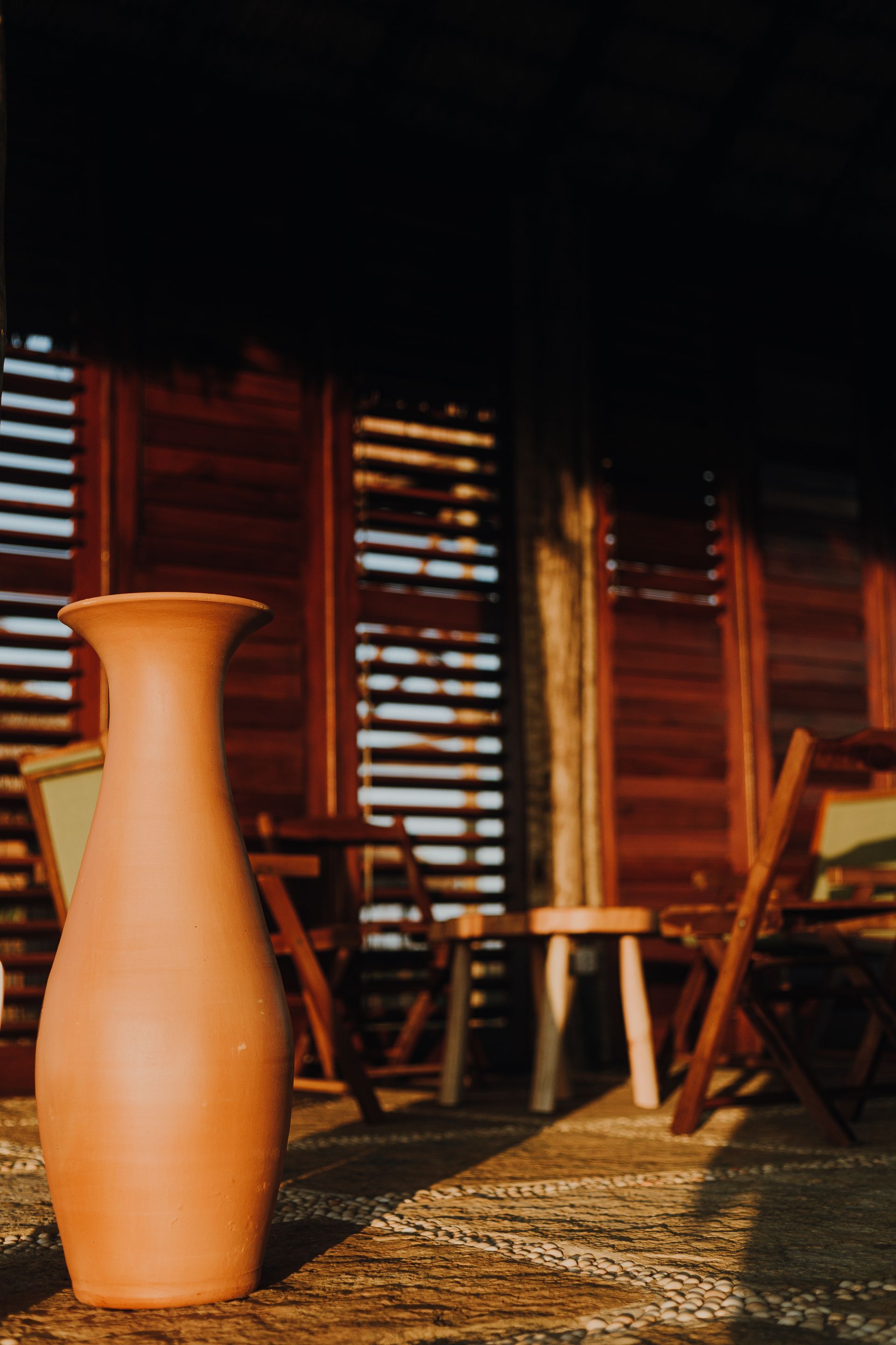 Vaso de terracota sobre uma superfície texturizada em frente a uma estrutura de madeira com venezianas abertas, uma mesa e cadeiras.
