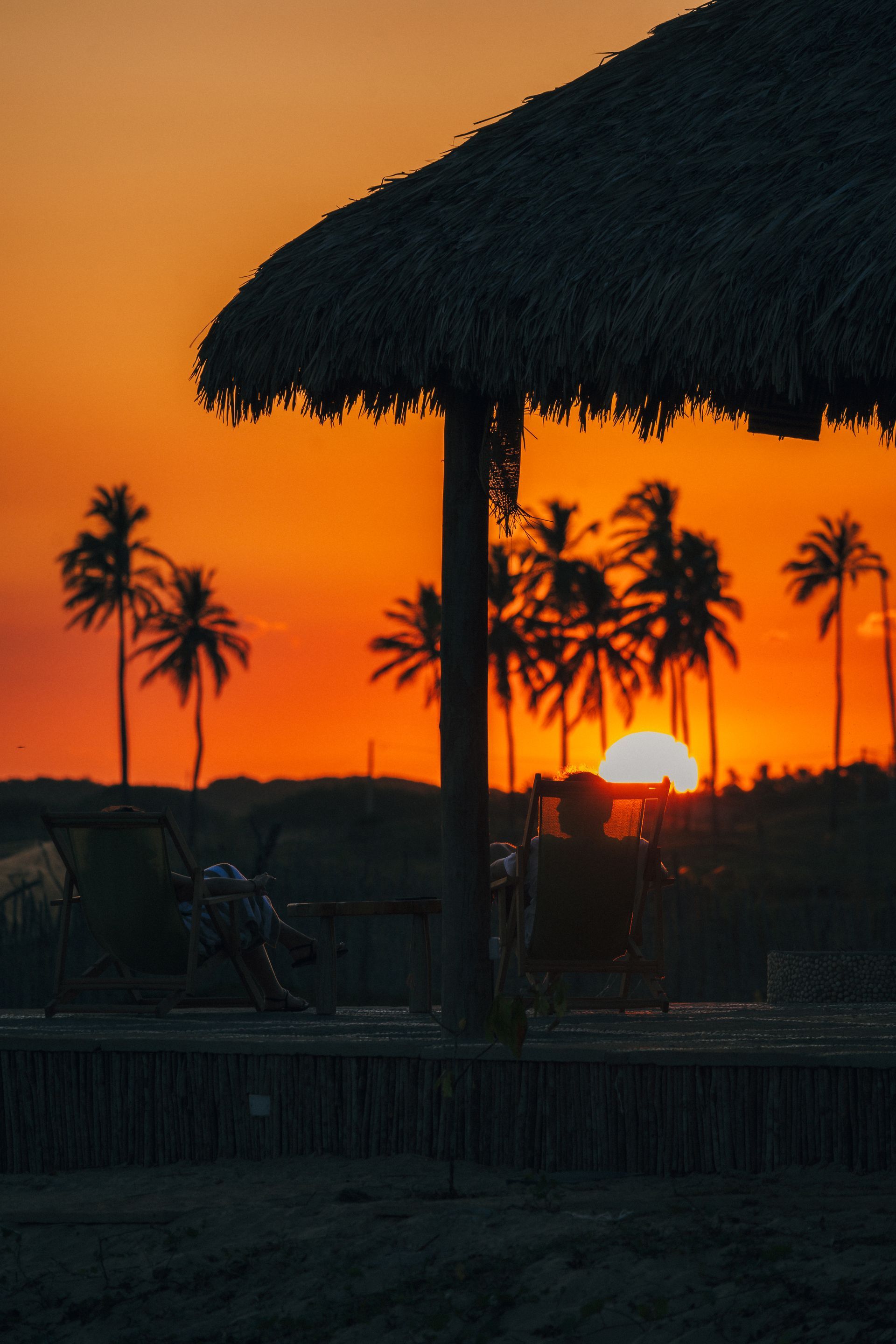 Silhueta de um abrigo de praia com telhado de palha e cadeiras contra um pôr do sol alaranjado vibrante com palmeiras ao fundo.