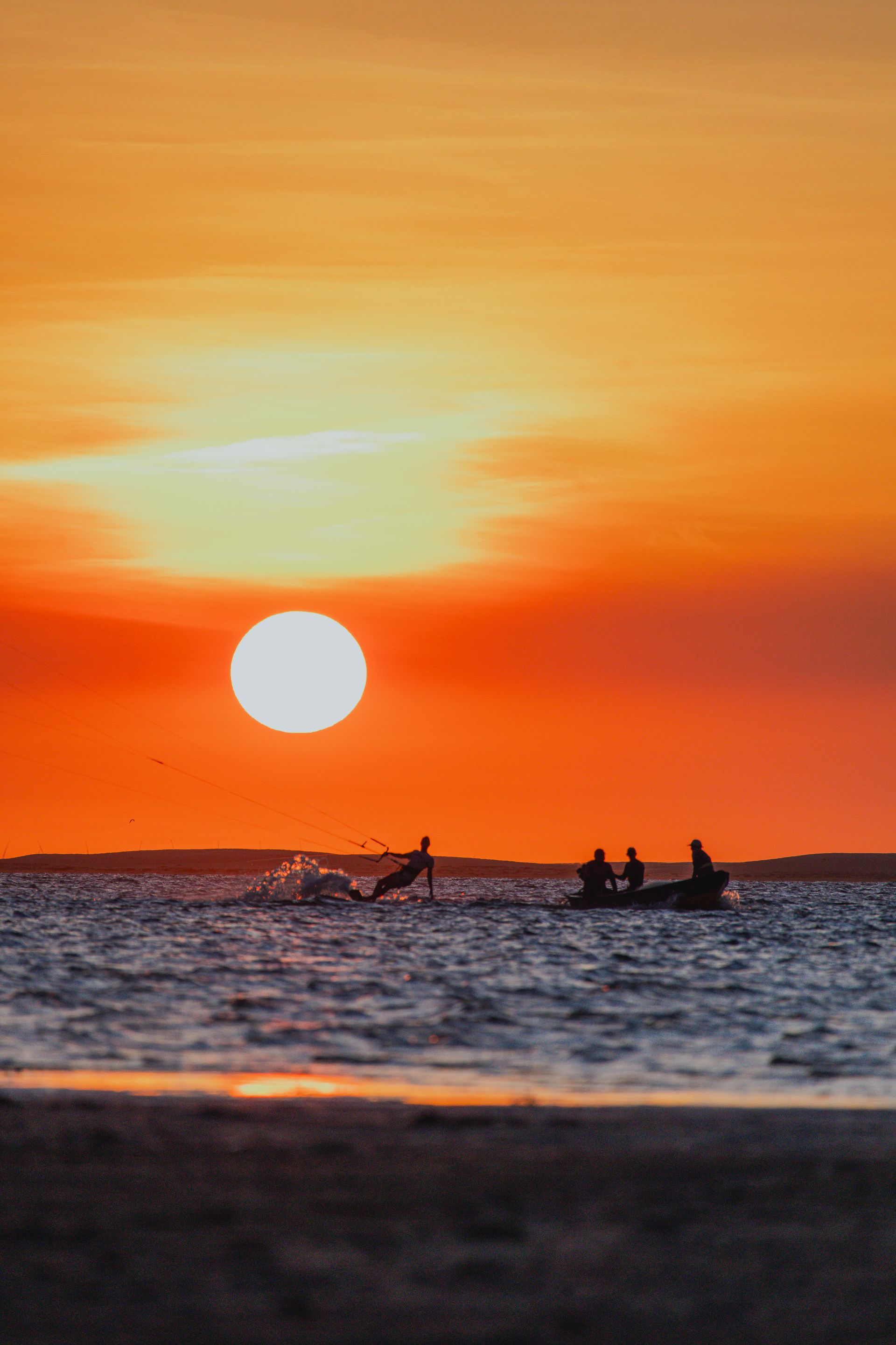 Pôr do sol sobre a água, um barco com figuras em silhueta. Céu alaranjado, sol próximo ao horizonte.
